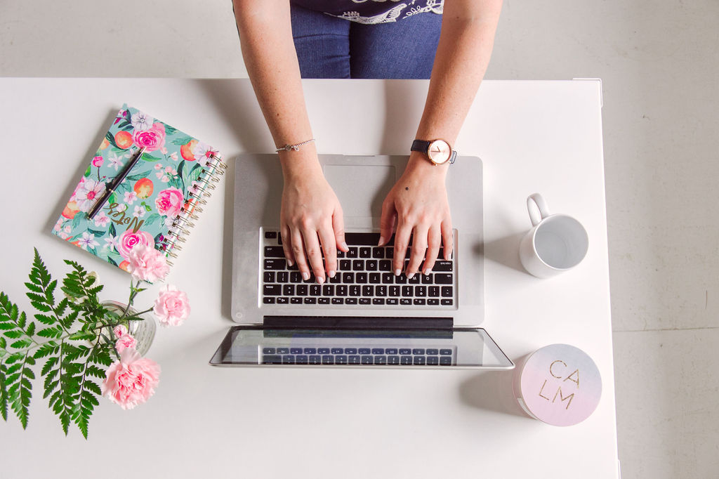 photo of woman typing on laptop computer with notebook and flowers on white desk